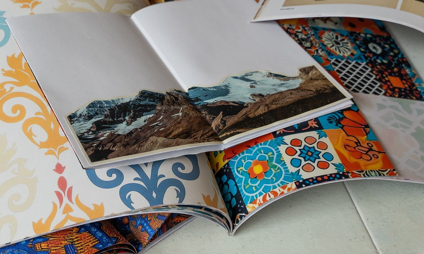 Two open zines on a table: a collage of mountains pasted across a blank spread, over pages of printed patterns